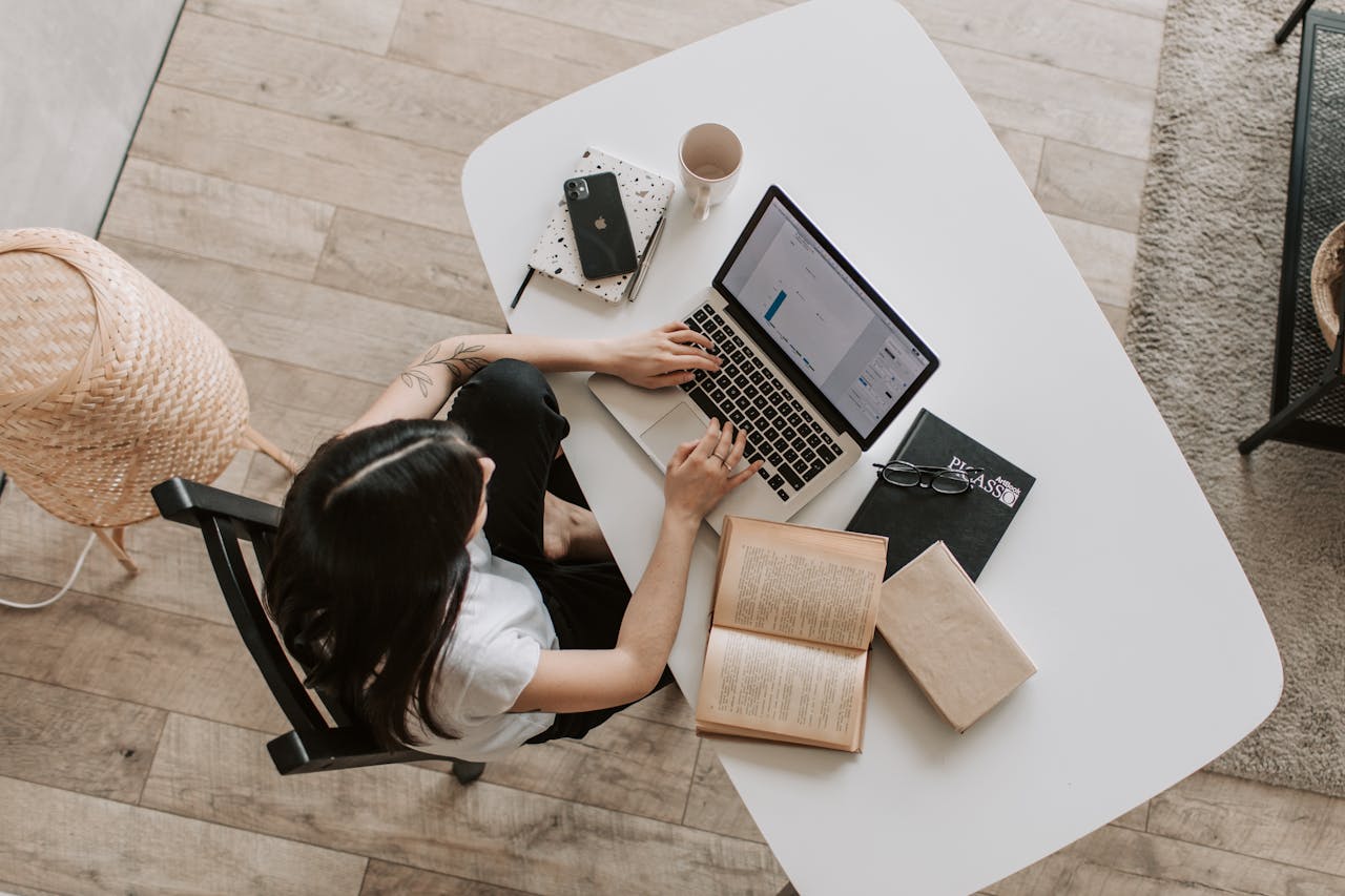 about-02 Overhead view of a woman using a laptop at a home desk, surrounded by books, a phone, and a cup.